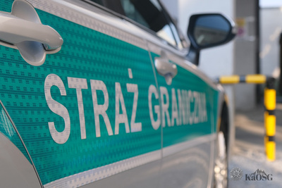 Bok srebrnego samochodu służbowego ze srebrnym napisem na zielonym tle straż graniczna w tle czarno-żółty słupek parkingowy