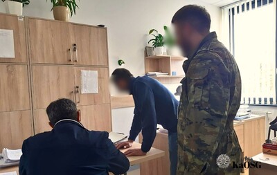 Na zdjęciu pomieszczenie biurowe, po lewej stronie siedzący na krześle mężczyzna prawdopodobnie podpisuje dokumenty. W środku obrazu pochylony inny mężczyzna opierający dłonie na blacie biurka przygląda się dokumentom. Po stronie prawej stojący mężczyzna w mundurze moro. W tle szafki biurowe.