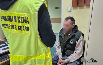 Na zdjęciu siedzący na krześle mężczyzna z zaplecionymi na kolanach dłońmi. Przed nim widoczny tyłem inny mężczyzna w czarnej bluzie i kamizelce odblaskowej z napisem Straż Graniczna.