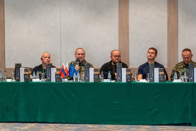 Uczestnicy szkolenia siedzą przy stole prezydialnym, jeden z nich przemawia do mikrofonu. Na stole flagi słowacji, polski i unijna.