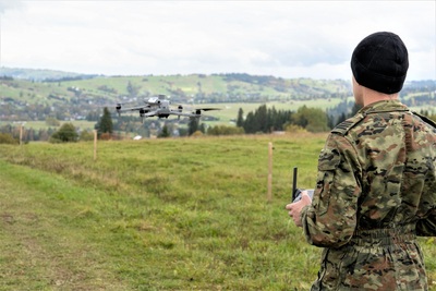 Funkcjonariusz w rękach trzyma w rękach kontroler drona. Przed nim w powietrzu unosi się szary dron. W tle zielona trawa i w oddali pagórki.