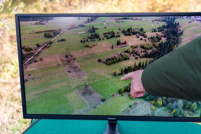 Telewizor pokazuje widok z góry z drona. Jeden z funkcjonariuszy pokazuje na monitorze palcem  miejsce pozostawienia odpadów, symulowane na potrzeby ćwiczeń terenowych.