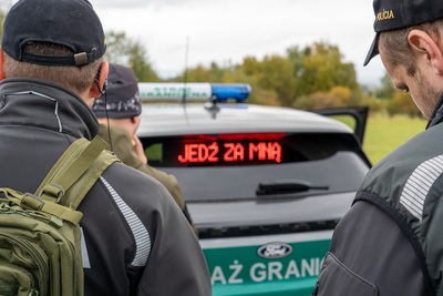Na pierwszym planie trzech funkcjonariuszy słowackiej policji stojących tyłem - widać ich głowy i ramiona. Przed nimi stoi zakupiony samochód oznakowany napisem Straż graniczna. Na podszybiu z tyłu samochodu wyświetla się napis  Jedź za mną.