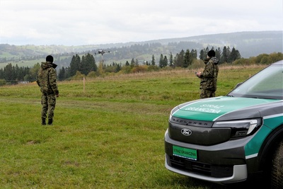 Dwóch funkcjonariuszy prowadzi lot dronem zakupionym w ramach projektu. Na pierwszym planie po prawej stronie przód samochodu z napisem straż graniczna. W tle pagórki.