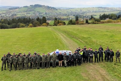Ujęcie z góry uczestników ćwiczeń terenowych. Za nimi stoi samochód zakupiony w ramach projektu. W tle drzewa i pagórki.