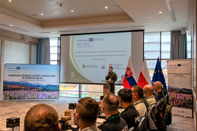 Uczestnicy projektu słuchają wystąpienia przewodniczącego projektu. Na monitorze wyświetla się prezentacja. Po bokach ekranu stoi ścinka i rolup projektowy.