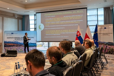 Uczestnicy projektu słuchają wystąpienia prelegenta. Na monitorze wyświetla się prezentacja. Po bokach ekranu stoi ścinka i rolup projektowy.