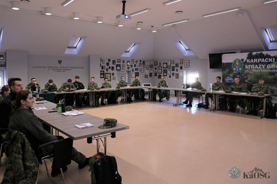 Nowo przyjęci funkcjonariusze siedzą przy stołach ułożonych w kształt litery u podczas prelekcji.