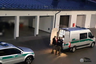 Na zdjęciu widać dwa srebrne pojazdy Straży Granicznej, do pojazdu po prawej stronie dwóch funkcjonariuszy stoi wraz z mężczyznom zatrzymanym, który będzie przewożony samochodem. W tle widać budynek w którym usytuowane są garaże z podniesionymi bramami.