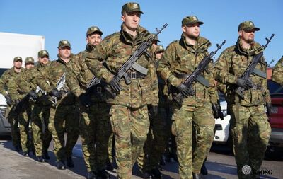 Szeroki plan przedstawiający kilkunastu żołnierzy lub funkcjonariuszy w mundurach maskujących typu moro, stojących w równym szeregu na baczność. Trzymają broń przewieszoną na ramieniu. Scena rozgrywa się na zewnątrz, w tle widoczne wysokie drzewa iglaste i liściaste oraz fragment budynku po prawej. Po lewej stronie widać zieloną flagę na maszcie.