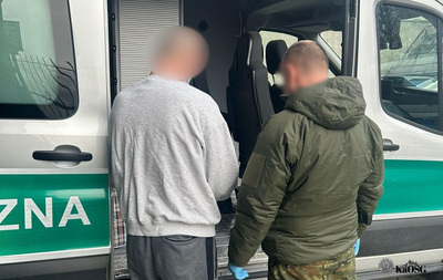 Na zdjęciu dwóch mężczyzn na tle fragmentu otwartego samochodu służbowego typu bus. Mężczyzna po prawej jest ubrany w spodnie moro i kurtkę koloru zielonego. Mężczyzna po lewej w bluzę dresową i spodnie dresowe.