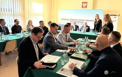 Kadr ze spotkania / konferencji. Widoczna jest baner Karpacki Oddział Straży Granicznej w Nowym Sączu oraz kilka osób przy stole z laptopami i notatkami.