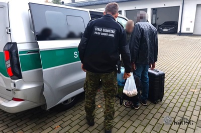 Na parkingu stoi bokiem z otwartymi przesuwnymi drzwiami samochód Straży Granicznej przy którym stoją dwaj funkcjonariusze oraz deportowany cudzoziemiec