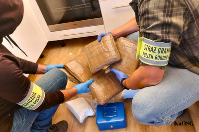 Na zdjęciu widać dwie osoby, które w pozycji kucznej sprawdzają woreczki z białym proszkiem. Na rękach mają założone niebieskie rękawiczki. Na ramionach mają opaski z napisem Straż Graniczna.