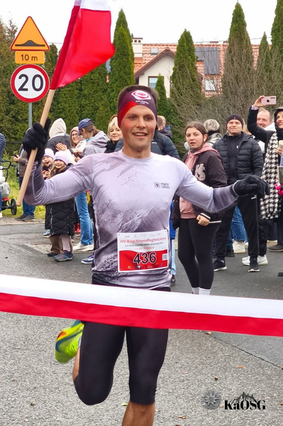 Na zdjęciu widać mężczyznę w stroju sportowym, który biegnie niosąc flagę biało-czerwoną. W tle widać innych ludzi.