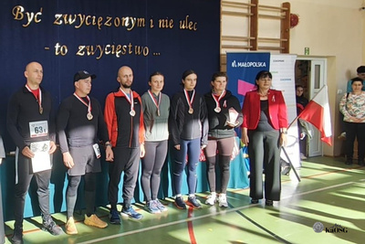 Na zdjęciu widać grupę ludzi, którzy stoją obok siebie. Zdjęcie zrobione na sali gimnastycznej w tle widać granatową tablicę z białymi napisami i częściowo zza nich widać drewniane drabinki.