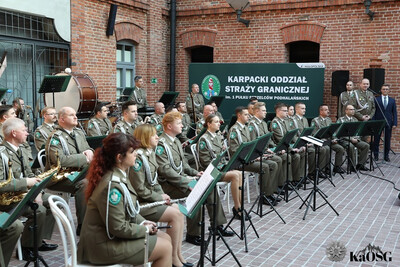 Orkiestra straży granicznej siedzi na krzesłach podczas koncertu, do mikrofonu przemawia generał, a w tle baner ciemnozielony z napisem karpacki oddział straży granicznej