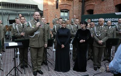 soliści koncertu - dwie kobiety jeden mężczyzna stoją w obecności generała, w tle siedzą muzycy orkiestry