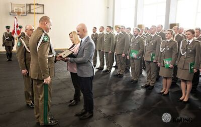 Hala sportowa. generał wręcza pracownikowi w garniturze odznakę kaosg na drewnianej tabliczce