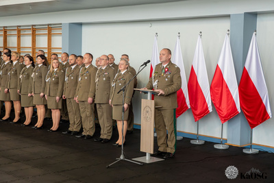 generał przemawia do mikrofonu. w tle kadra kierownicza oddziału stojąca w dwuszeregu. po prawej biało-czerwone falgi