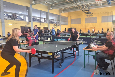 Na zdjęciu widać halę sportową z rozłożonymi stołami do gry w ping ponga. Dwie zawodniczki grają, po prawej stronie widać mężczyznę, który im sędziuje.