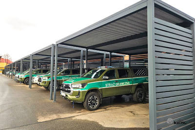 Na zdjęciu widać popielate wiaty, pod którymi stoją samochody srebrne z zielonymi napisami.