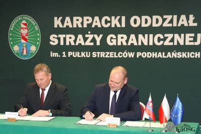 Na zdjęciu widać dwóch mężczyzn którzy podpisują dokumenty. W tle widać po lewej stronie zielone logo, na środku widać duży napis karpacki Oddział Straży Granicznej.