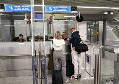 Odprawa paszportowa na lotnisku. Dwie osoby z bagażem podręcznym rozmawiają z funkcjonariuszem Straży Granicznej przy boksie kontrolnym. Nad stanowiskiem widoczna jest niebieska tablica informacyjna „Kontrola Graniczna / Border Control”.