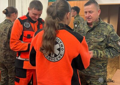 Grupa osób w mundurach Straży Granicznej oraz strojach ratowniczych uśmiecha się i trzyma w dłoniach białe opłatki wigilijne. Na pierwszym planie po lewej kobieta w mundurze z naszywką formacji, po prawej uśmiechnięty ratownik medyczny. Atmosfera jest radosna i świąteczna.