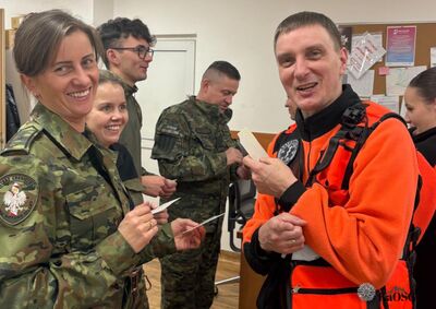 Zbliżenie na rozmowę dwóch mężczyzn i kobiety. Po prawej mężczyzna w mundurze Straży Granicznej z widocznymi pagonami, po lewej mężczyzna w kamizelce ratownika medycznego. Tyłem do obiektywu stoi kobieta w pomarańczowej bluzie z dużym emblematem „Ratownictwo Medyczne” i gwiazdą życia na plecach.