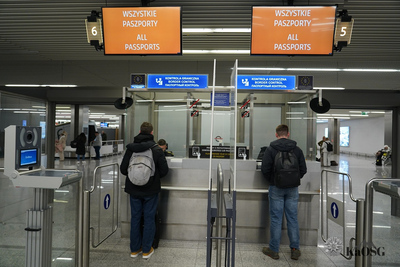 Na zdjęciu widać kontrolę graniczną na lotnisku Do stanowiska kontrolerskiego przy okienkach stoi dwóch mężczyzn. Funkcjonariusze SG kontrolują dokumenty podróży. W tle widać halę na lotnisku.