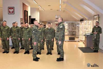 Funkcjonariusz melduje gotowość oddziału komendantowi. Pozostali funkcjonariusze stoją w postawie zasadniczej w sali tradycji.