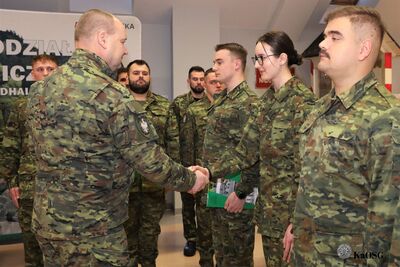 Zbliżenie na moment uścisku dłoni między komendantem a nową funkcjonariuszką. Funkcjonariusze trzymają zielone foldery pamiątkowe.