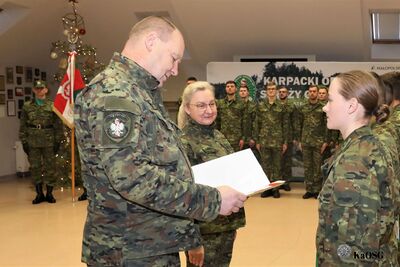 Komendant jednostki w kamuflażu polowym wręcza dokument młodej funkcjonariuszce. W tle widać poczet sztandarowy oraz udekorowaną choinkę.