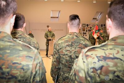 Widok zza pleców funkcjonariuszy na komendanta przemawiającego przy mównicy. Po prawej stronie stoi poczet sztandarowy ze sztandarem Karpackiego Oddziału Straży Granicznej.