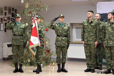 Poczet sztandarowy Straży Granicznej w postawie "Prezentuj broń" (oddawanie honorów). W tle znajduje się choinka i ściana z pamiątkowymi ryngrafami.