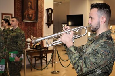 Profil funkcjonariusza Straży Granicznej grającego na trąbce podczas oficjalnej części uroczystości. W tle widoczne są eksponaty muzealne i portret w ramie.