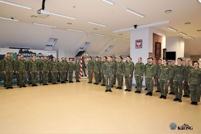 Szeroki kadr na salę tradycji, w której w dwóch szeregach stoją funkcjonariusze i funkcjonariuszki w umundurowaniu polowym. Na ścianach widoczne są symbole narodowe i godło Polski.