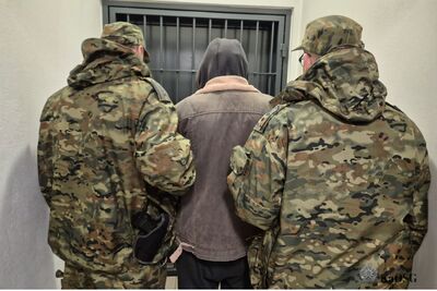 Na zdjęciu dwóch mężczyzn w umundurowaniu moro trzymają pod rękę innego mężczyznę ubranego w kurtkę, z kapturem na głowie. Przed nimi drzwi z kratą.