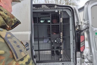 Na zdjęciu mężczyzna siedzący w samochodzie typu więźniarka. Po lewej stronie fragment mężczyzny ubranego w umundurowanie moro.