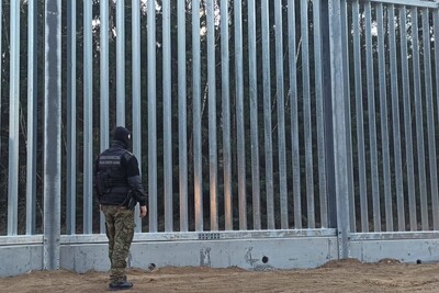 Funkcjonariusz Straży Granicznej w pełnym umundurowaniu i kominiarce stoi na straży przy wysokiej, stalowej barierze granicznej na tle lasu.