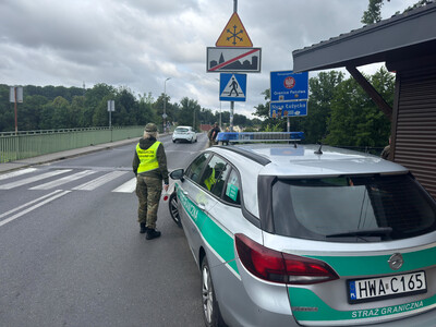 Funkcjonariuszka Straży Granicznej w odblaskowej kamizelce stoi przy radiowozie na drodze dojazdowej do mostu granicznego. W tle widoczne polskie znaki drogowe: "Granica Państwa" oraz "Nysa Łużycka".