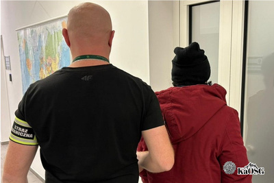 zatrzymany cudzoziemiec prowadzony jest przez nieumundurowanego funkcjonariusza sg z odblaskową opaską na ramieniu z napisem straż graniczna