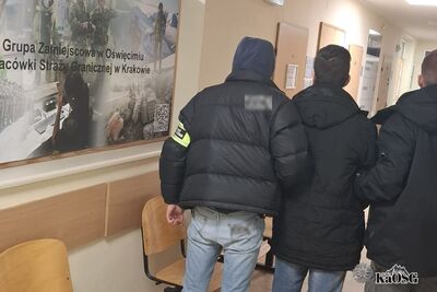 Na zdjęciu dwóch mężczyzn w cywilnych ubraniach idących korytarzem. Na rękawie jednego z nich (po lewej) opaska z napisem Straż Graniczna. Prowadzą pod rękę trzeciego mężczyznę będącego prawdopodobnie osobą zatrzymaną. Po lewej stronie zdjęcia tablica na ścianie z napisem Grupa Zamiejscowa w Oświęcimiu, Placówka Straży Granicznej w Krakowie.