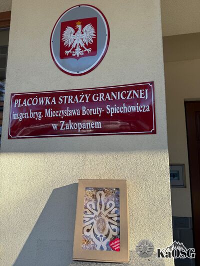 Na zdjęciu w górnej części tabliczka z napisem Placówka Straży Granicznej im. gen. bryg. Mieczysława Boruty - Spiechowicza w Zakopanem. Nad nią blaszana tabliczka z godłem Polski. Poniżej gadżet w postaci drewnianej parzenicy w ozdobnym opakowaniu.