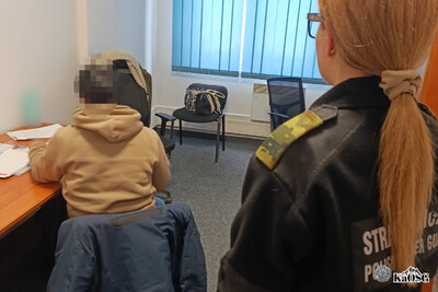 Cudzoziemiec siedzi przy biurku, pilnuje go funkcjonariuszka straży granicznej