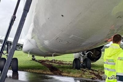 Na zdjęciu fragment samolotu pasażerskiego, który zjechał najprawdopodobniej z pasa startowego. Po prawej stronie mężczyzna ubrany w odblaskową kamizelkę z napisem Airport Services.