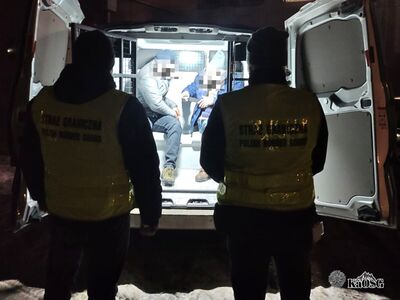 Na zdjęciu dwaj mężczyźni w kamizelkach odblaskowych, stoją przodem do samochodu typu więźniarka. Auto ma otwarte tylne drzwi a wewnątrz znajduje się dwóch mężczyzn siedzących, prawdopodobnie osoby zatrzymane.