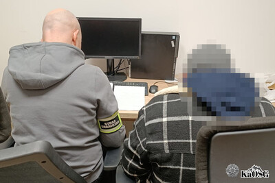 Zatrzymany cudzoziemiec podpisuje na biurku dokumenty. Towarzyszy mu funkcjonariusz w stroju cywilnym z opaską odblaskową na ramieniu z napisem straż graniczna