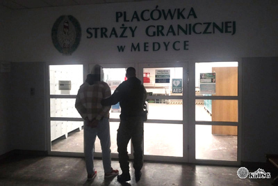 Na zdjęciu dwóch mężczyzn w mundurach trzyma osobę zatrzymaną i czekają aż wsiądzie do samochodu do przewożenia osób zatrzymanych.